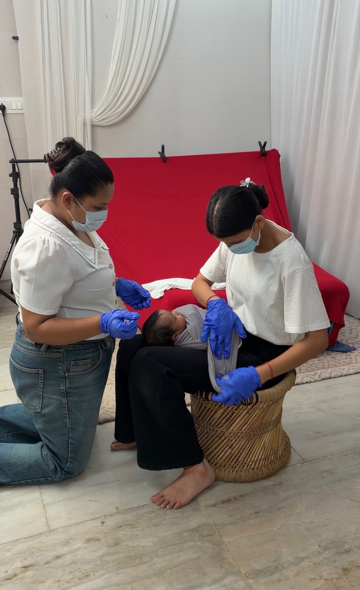 Two team members in masks and blue gloves, hair tied back, wrapping a newborn in a grey stretch wrap on one team member’s lap in the studio.