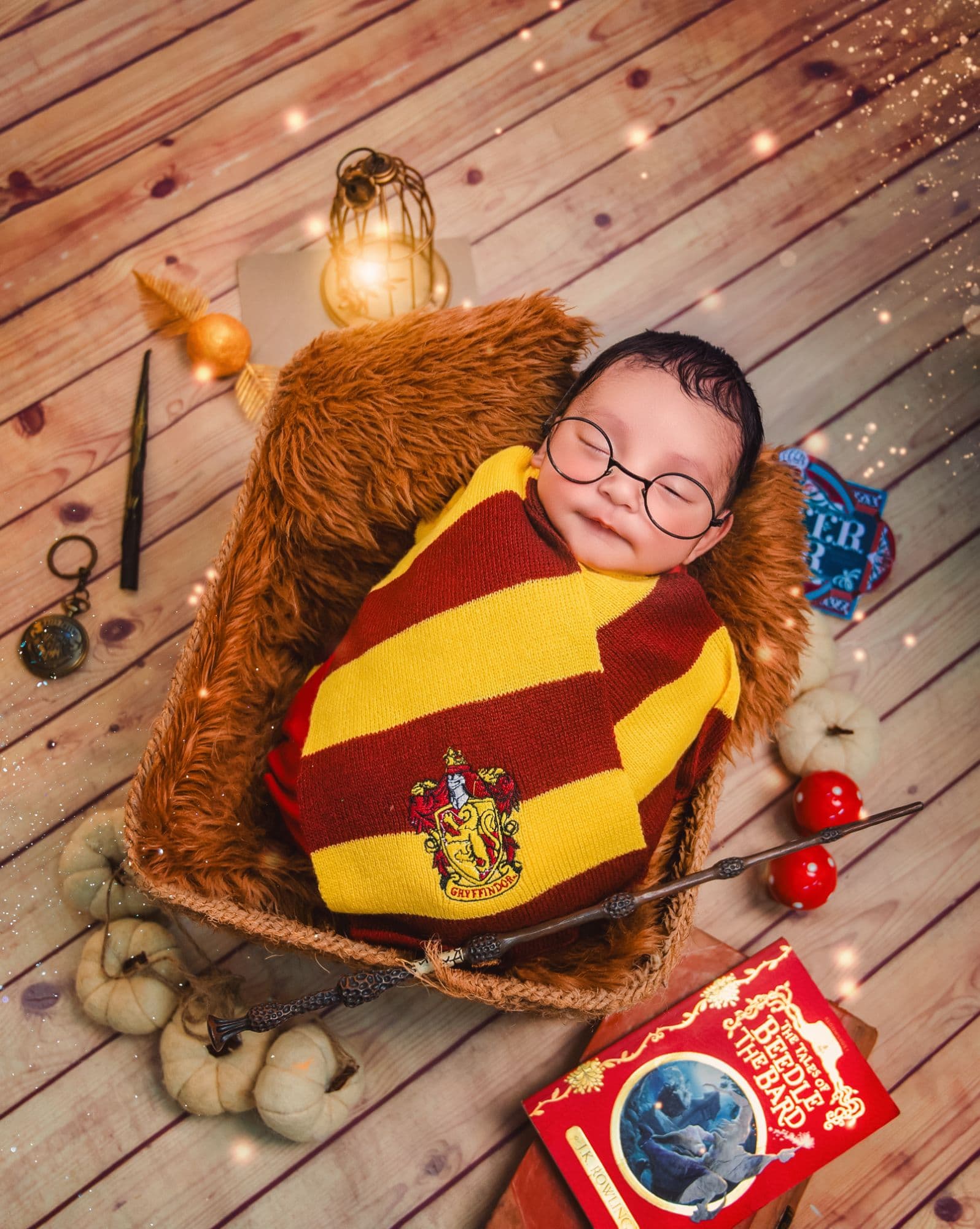 Sleeping newborn swaddled in a maroon and gold striped knit scarf, wearing round prop glasses, resting in a fur-lined basket surrounded by storybook props.